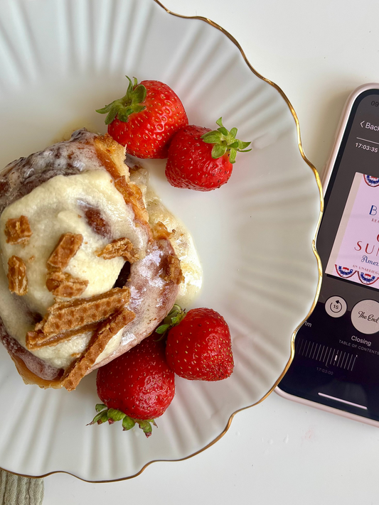 Cinnamon roll with icing and strawberries on a plate next to a smartphone displaying an audiobook cover.